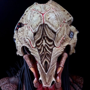 May include: A detailed, hand-painted bust of a Predator-like creature with a skull-like head, red and white markings, and black dreadlocks. The bust is made of a light brown material and has a textured surface.