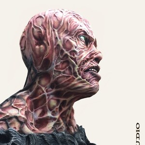 May include: A detailed bust sculpture of a creature with red, yellow, and black textured skin. The creature has a human-like face with a wide open mouth and visible teeth. The sculpture is made of a dark grey material and has a textured surface.