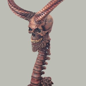 May include: A wooden sculpture of a skull with horns and a spine. The skull is painted in shades of brown and red, and the horns are curled.