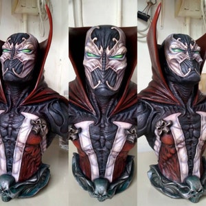May include: A detailed bust of the superhero Spawn, painted in red, black and white. The bust features a sculpted, detailed face with green eyes and a cape.