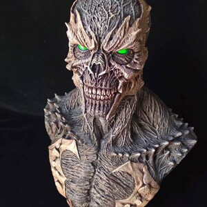 May include: A detailed bust of a demonic figure with glowing green eyes. The sculpture has a textured, weathered appearance with sharp, spiky details around the head and shoulders. The bust is a custom statue from Buster Studio.
