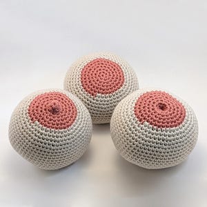 May include: Three handmade crochet spheres, each featuring a light beige base with a circular, rose-colored center. The spheres are arranged on a white surface, showcasing the detailed texture of the crochet work. The design is simple and artistic.