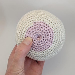 May include: A crocheted sphere in off-white with a central pink circle. The sphere is held in a hand, showcasing the texture and color contrast. The handmade item is likely a decorative or functional piece.
