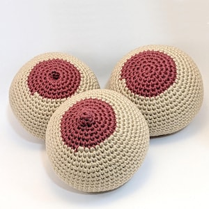 May include: Three handmade crochet spheres with a beige exterior and a maroon central circle, resembling a breast. The spheres are arranged in a triangular formation against a white background. The crochet work is detailed and textured.