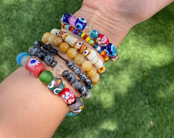 May include: A stack of colourful beaded bracelets with various charms, including evil eye beads, gemstone beads, and wooden beads. The bracelets are worn on a person's wrist.