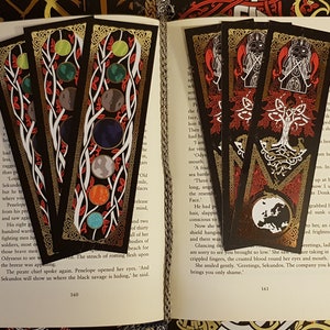 Yggdrasil Gold Foil Double-Sided Bookmarks strewn over the pages of an open book with a Nordic style depiction of the life tree and the world in the colours of black, red, white and gold on one side and the planets of the solar system on the other.