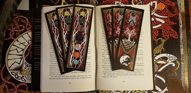 Yggdrasil Gold Foil Double-Sided Bookmarks strewn over the pages of an open book with a Nordic style depiction of the life tree and the world in the colours of black, red, white and gold on one side and the planets of the solar system on the other.