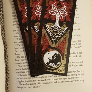 Bookmarks strewn over an open page of a book. Yggdrasil Gold Foil Double-Sided Bookmark with a Nordic style depiction of the life tree and the world in the colours of black, red, white and gold on one side.