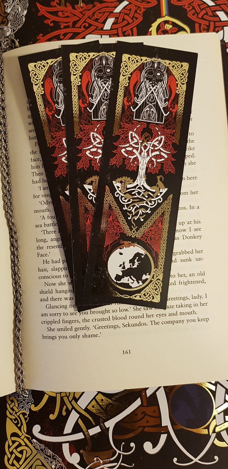 Bookmarks strewn over an open page of a book. Yggdrasil Gold Foil Double-Sided Bookmark with a Nordic style depiction of the life tree and the world in the colours of black, red, white and gold on one side.