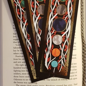 Bookmarks strewn over an open page of a book. Yggdrasil Gold Foil Double-Sided Bookmark with close up of the side with planets of the solar system on a background in the colours of black, red, white and gold.