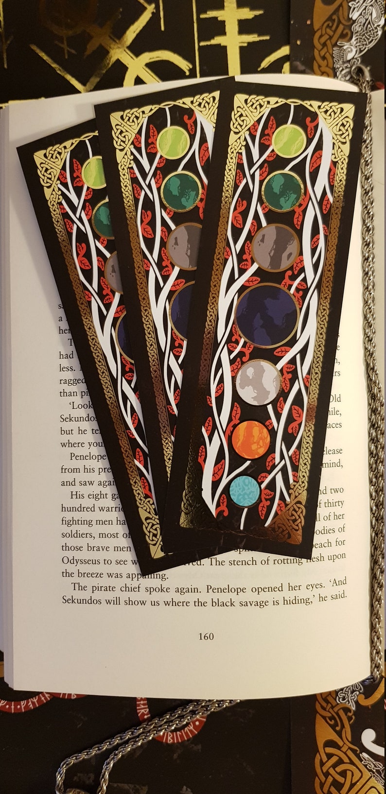 Bookmarks strewn over an open page of a book. Yggdrasil Gold Foil Double-Sided Bookmark with close up of the side with planets of the solar system on a background in the colours of black, red, white and gold.