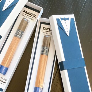 Puede incluir: Cajas blancas con dise&ntilde;os de esmoquin azules y cigarros. Cada caja contiene un cigarro con cuerpo de madera y punta azul. El texto en las cajas dice "PARKER, Time to Suit Up!" y "TAYLOR, Time to Suit Up!" y "Will you be my, GROOMSMAN?"