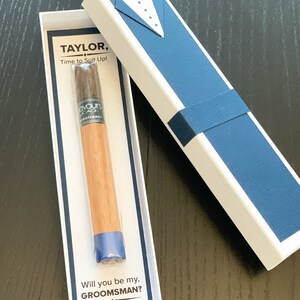 Puede incluir: Un puro en una caja blanca con un dise&ntilde;o de esmoquin azul. La caja dice "TAYLOR, Time to Suit Up!" y "Will you be my, GROOMSMAN? JON."