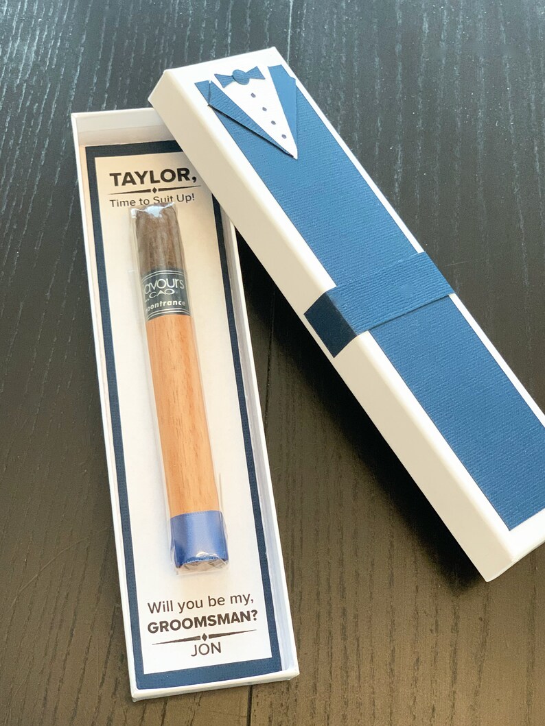 Puede incluir: Un puro en una caja blanca con un dise&ntilde;o de esmoquin azul. La caja dice "TAYLOR, Time to Suit Up!" y "Will you be my, GROOMSMAN? JON."