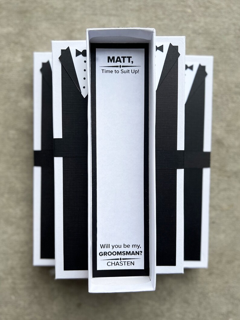 Puede incluir: Tres cajas blancas con dise&ntilde;os de esmoquin negros y el texto "MATT, Time to Suit Up!" y "Will you be my, GROOMSMAN? CHASTEN". Las cajas est&aacute;n atadas con cintas negras, perfectas para una boda.
