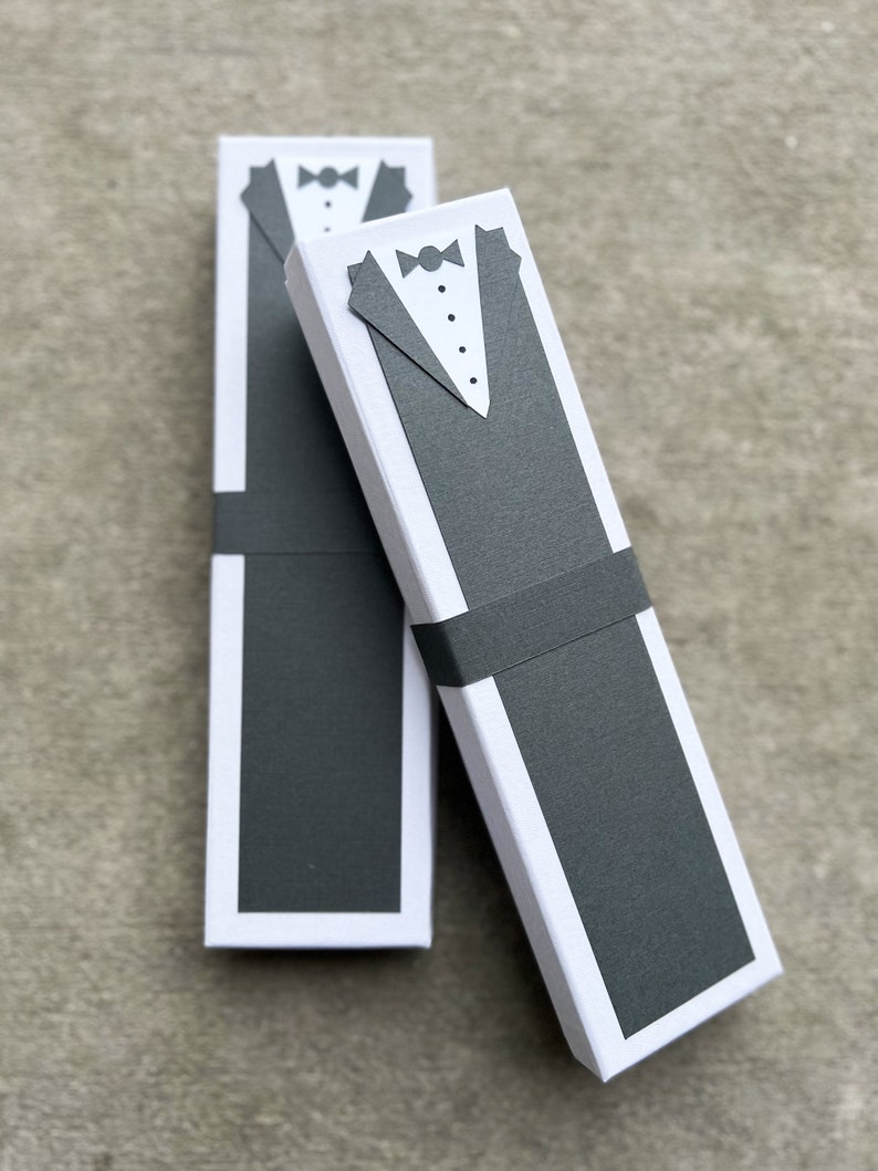 Puede incluir: Dos cajas de regalo rectangulares blancas con un dise&ntilde;o de esmoquin. Las cajas tienen un dise&ntilde;o de chaqueta de esmoquin negra y pajarita en la parte delantera. Una banda negra envuelve el centro de cada caja. Las cajas est&aacute;n sobre una superficie gris.