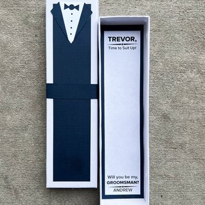 Puede incluir: Una caja blanca con un dise&ntilde;o de esmoquin azul marino en la parte delantera. La caja est&aacute; abierta, revelando una tarjeta con el texto "TREVOR, Time to Suit Up!" y "Will you be my, GROOMSMAN?" con el nombre "ANDREW" debajo.