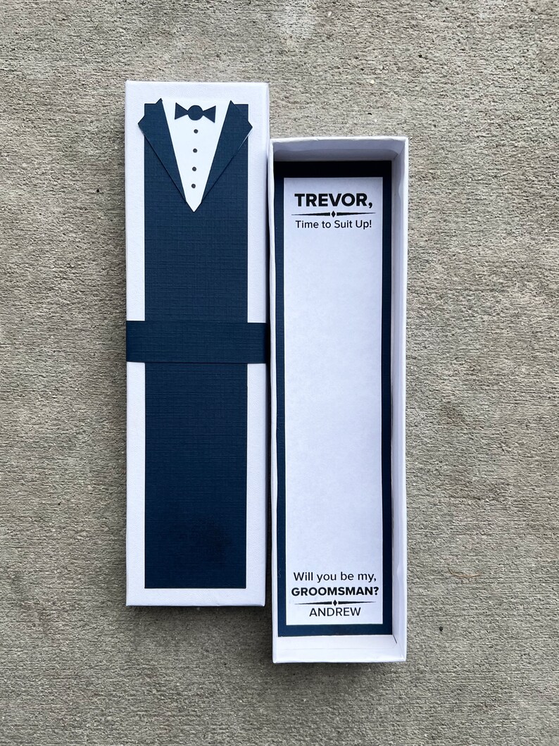 Puede incluir: Una caja blanca con un dise&ntilde;o de esmoquin azul marino en la parte delantera. La caja est&aacute; abierta, revelando una tarjeta con el texto "TREVOR, Time to Suit Up!" y "Will you be my, GROOMSMAN?" con el nombre "ANDREW" debajo.