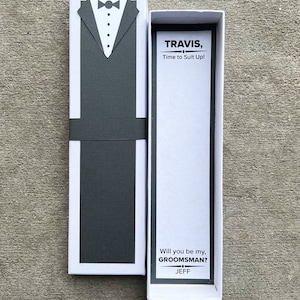 Puede incluir: Una caja de regalo blanca con un dise&ntilde;o de esmoquin gris en la parte delantera. La caja est&aacute; abierta, revelando el texto "TRAVIS, Time to Suit Up!" y "Will you be my, GROOMSMAN? JEFF".