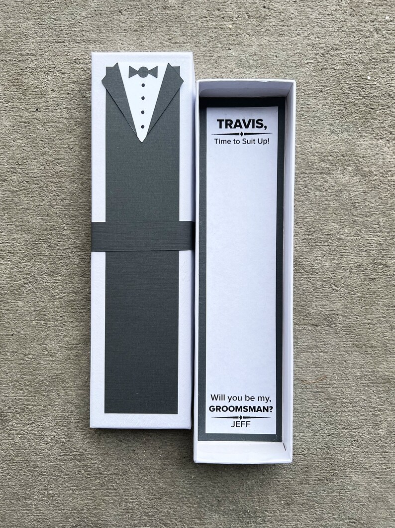 Puede incluir: Una caja de regalo blanca con un dise&ntilde;o de esmoquin gris en la parte delantera. La caja est&aacute; abierta, revelando el texto "TRAVIS, Time to Suit Up!" y "Will you be my, GROOMSMAN? JEFF".
