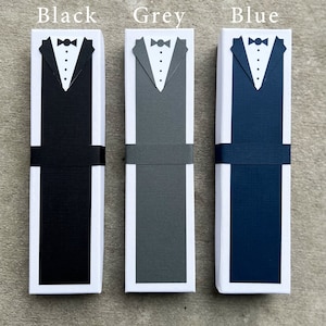 Puede incluir: Tres cajas de regalo rectangulares, cada una con un dise&ntilde;o de esmoquin. Las cajas son negras, grises y azules, con camisas blancas, pajaritas negras y una banda negra. El texto "Black", "Grey" y "Blue" est&aacute; encima de cada caja.