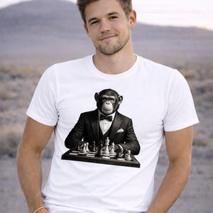 Può includere: T-shirt bianco con un'immagine in bianco e nero di uno scimpanzé in smoking che gioca a scacchi. Lo scimpanzé è seduto davanti a una scacchiera. La persona che indossa la maglietta sorride.