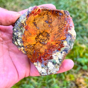 May include: A close-up of a colorful agate stone with a variety of brown, orange, red, white, and green patterns. The stone is being held in a hand.