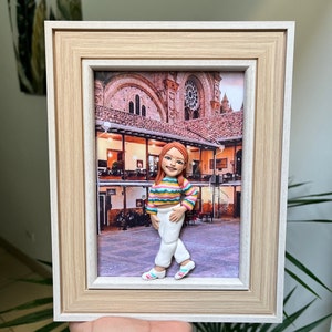 K&ouml;nnte beinhalten: Ein gerahmtes Bild einer Tonfigur einer Person, die vor einem Geb&auml;ude steht. Die Figur tr&auml;gt ein gestreiftes Hemd, eine wei&szlig;e Hose und rosa Schuhe. Das Geb&auml;ude ist eine Kirche mit einem gro&szlig;en Fenster und einem Ziegeldach.