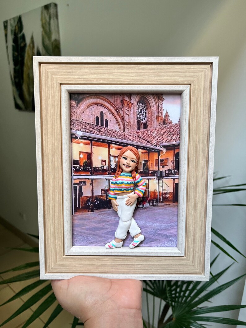 K&ouml;nnte beinhalten: Ein gerahmtes Bild einer Tonfigur einer Person, die vor einem Geb&auml;ude steht. Die Figur tr&auml;gt ein gestreiftes Hemd, eine wei&szlig;e Hose und rosa Schuhe. Das Geb&auml;ude ist eine Kirche mit einem gro&szlig;en Fenster und einem Ziegeldach.