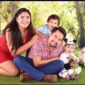 K&ouml;nnte beinhalten: Eine vierk&ouml;pfige Familie posiert f&uuml;r ein Foto in einem gr&uuml;nen Grasbereich mit einem Hintergrund aus B&auml;umen. Die Eltern sitzen auf dem Boden, w&auml;hrend die beiden Kinder stehen. Der Vater tr&auml;gt ein rot-wei&szlig; kariertes Hemd, blaue Jeans und braune Schuhe. Die Mutter tr&auml;gt ein rotes Kleid. Das &auml;ltere Kind tr&auml;gt ein blau-wei&szlig; kariertes Hemd. Das j&uuml;ngere Kind tr&auml;gt einen wei&szlig;en Strampler mit einer Panda-M&uuml;tze.