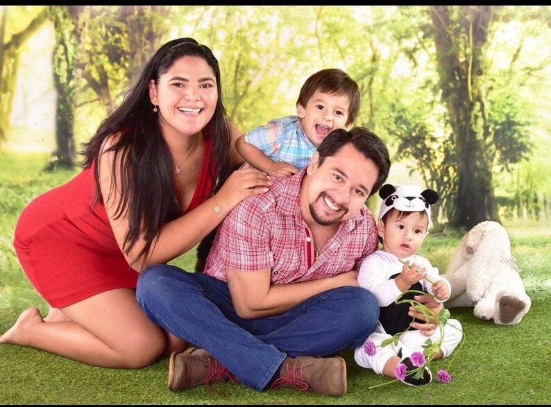 K&ouml;nnte beinhalten: Eine vierk&ouml;pfige Familie posiert f&uuml;r ein Foto in einem gr&uuml;nen Grasbereich mit einem Hintergrund aus B&auml;umen. Die Eltern sitzen auf dem Boden, w&auml;hrend die beiden Kinder stehen. Der Vater tr&auml;gt ein rot-wei&szlig; kariertes Hemd, blaue Jeans und braune Schuhe. Die Mutter tr&auml;gt ein rotes Kleid. Das &auml;ltere Kind tr&auml;gt ein blau-wei&szlig; kariertes Hemd. Das j&uuml;ngere Kind tr&auml;gt einen wei&szlig;en Strampler mit einer Panda-M&uuml;tze.