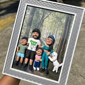 K&ouml;nnte beinhalten: Ein gerahmtes Foto einer vierk&ouml;pfigen Familie mit einem Hund. Die Familie besteht aus Tonfiguren und sie stehen in einer Waldkulisse. Der Vater tr&auml;gt ein gr&uuml;nes Hemd mit der Aufschrift "Green Day". Die Mutter tr&auml;gt ein blaues Hemd und eine schwarze Hose. Die beiden Kinder tragen blaue Hemden und Shorts. Der Hund ist wei&szlig; und flauschig.