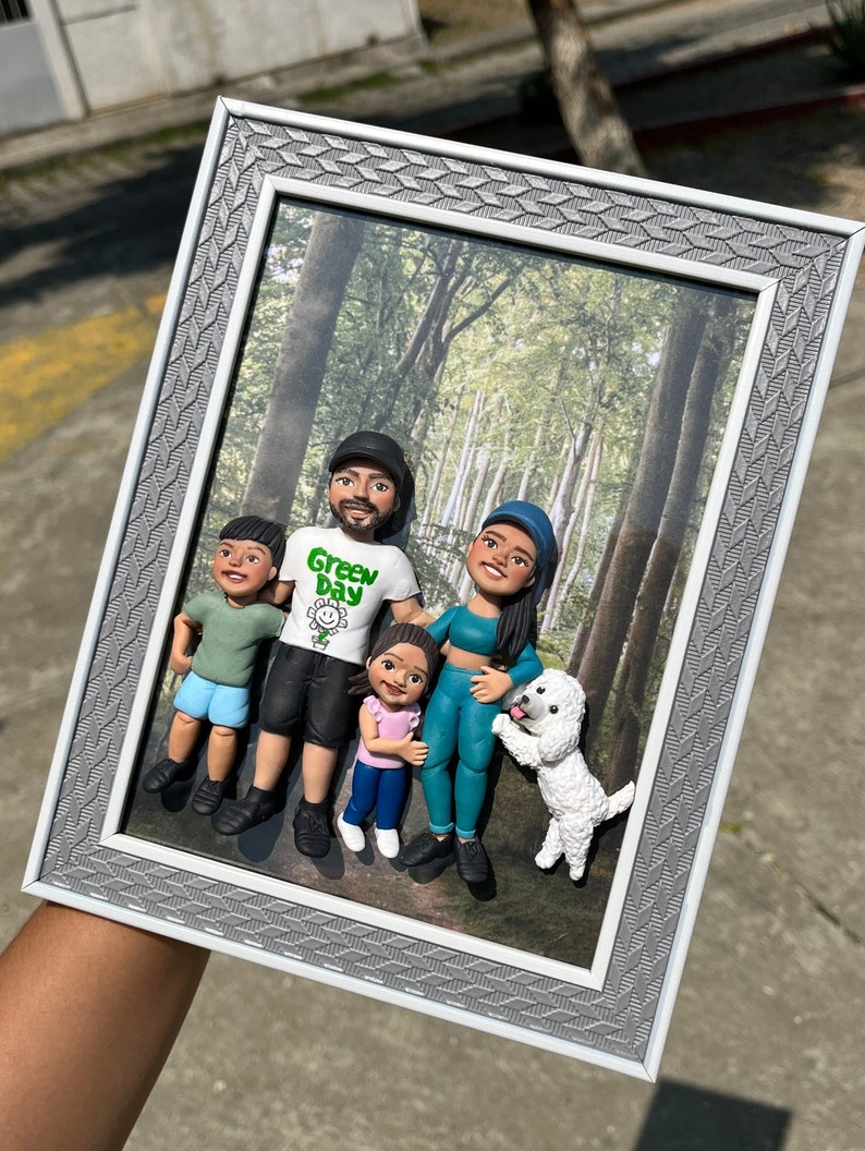 K&ouml;nnte beinhalten: Ein gerahmtes Foto einer vierk&ouml;pfigen Familie mit einem Hund. Die Familie besteht aus Tonfiguren und sie stehen in einer Waldkulisse. Der Vater tr&auml;gt ein gr&uuml;nes Hemd mit der Aufschrift "Green Day". Die Mutter tr&auml;gt ein blaues Hemd und eine schwarze Hose. Die beiden Kinder tragen blaue Hemden und Shorts. Der Hund ist wei&szlig; und flauschig.