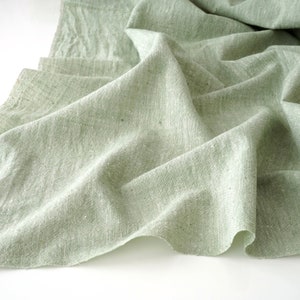 May include: A close-up of a light green linen fabric with a textured weave. The fabric is draped and folded, creating soft shadows and highlights. The colour is a muted, natural tone, suggesting a rustic or vintage aesthetic. The fabric appears to be lightweight and breathable.