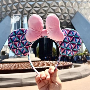 May include: Pink velvet bow and ears headband with a geometric pattern. The ears feature a colourful geometric design in shades of blue, pink, and purple. The headband is held up in front of a large, silver geodesic sphere.