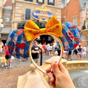 Op de afbeelding: Een haarband met twee ronde oren in een blauw-wit ruitpatroon, versierd met Ratatouille-karakterafbeeldingen en de tekst Gusteau's. Tussen de oren zit een felgele strik met een patroon van gaten zoals in kaas.