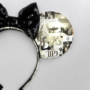 May include: Black and white patterned mouse ears with a black bow and a collage of images featuring Taylor Swift. The text "The Tortured Lets Department" and "But Daddy I Love Him" are visible on the ears. The text "T.S. Taylor Swift" is also visible.
