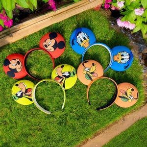 May include: Six pairs of character-themed headband ears. The ears are made of fabric and feature Mickey Mouse, Donald Duck, and Goofy. The headbands are white.
