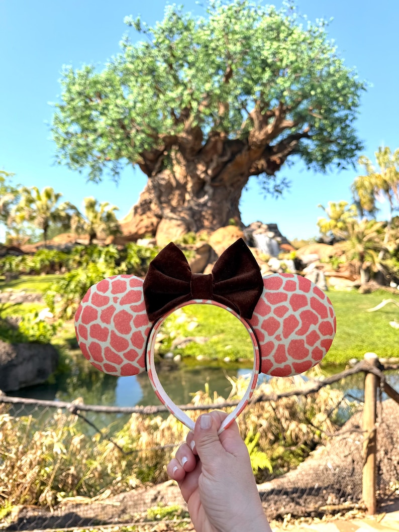 K&ouml;nnte beinhalten: Ein Stirnband mit Ohren im Maus-Design mit Giraffenmuster. Die Ohren sind korallenfarben mit wei&szlig;em Muster, die Schleife ist braun. Das Stirnband ist wei&szlig; mit einem korallenfarbenen und wei&szlig;en Muster. Im Hintergrund ein Park mit einem gro&szlig;en Baum.