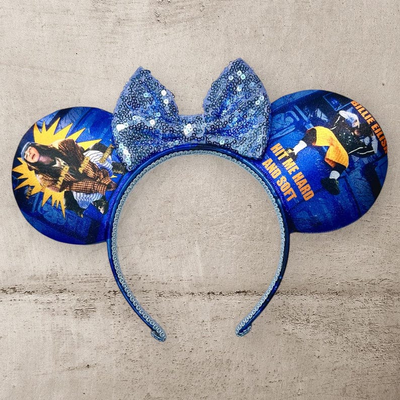 May include: Blue and yellow mouse ears with a blue bow. The ears feature a photo of Billie Eilish with the text "Hit Me Hard And Soft".