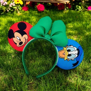 May include: Green headband with a red Mickey Mouse ear and a blue Donald Duck ear. The headband has a green bow in the center.