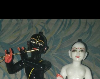 May include: Two marble statues of Hindu deities, one black and one white, standing on separate bases. The black statue is playing a flute, while the white statue has its hands raised in a gesture of prayer. The statues are both about 10 inches tall.
