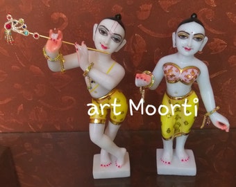 May include: White marble statue of Radha Krishna, a Hindu deity couple, with intricate details and colorful accents. The statue depicts Radha holding a string of beads and Krishna playing a flute.