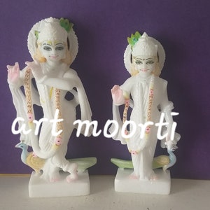 Puede incluir: Dos estatuas blancas de m&aacute;rmol de deidades hind&uacute;es, cada una sosteniendo una pluma de pavo real y luciendo joyas elaboradas. Las estatuas est&aacute;n de pie sobre bases blancas.