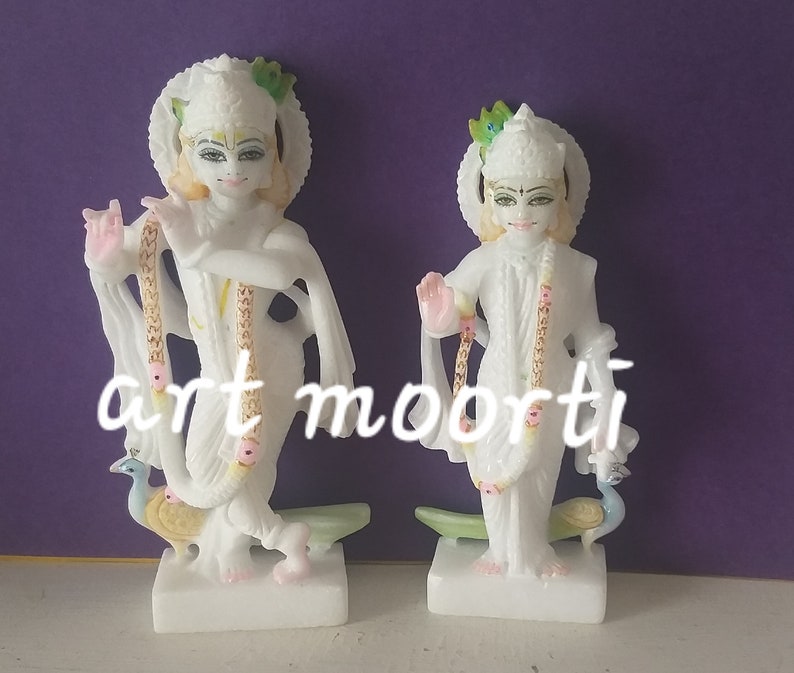 Puede incluir: Dos estatuas blancas de m&aacute;rmol de deidades hind&uacute;es, cada una sosteniendo una pluma de pavo real y luciendo joyas elaboradas. Las estatuas est&aacute;n de pie sobre bases blancas.