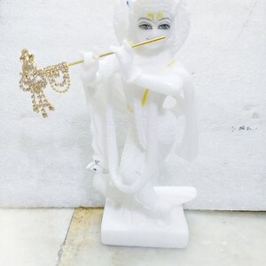 Puede incluir: Estatua de m&aacute;rmol blanco de una deidad hind&uacute; tocando la flauta. La estatua est&aacute; detallada con pintura dorada y tiene una pluma de pavo real en la cabeza.