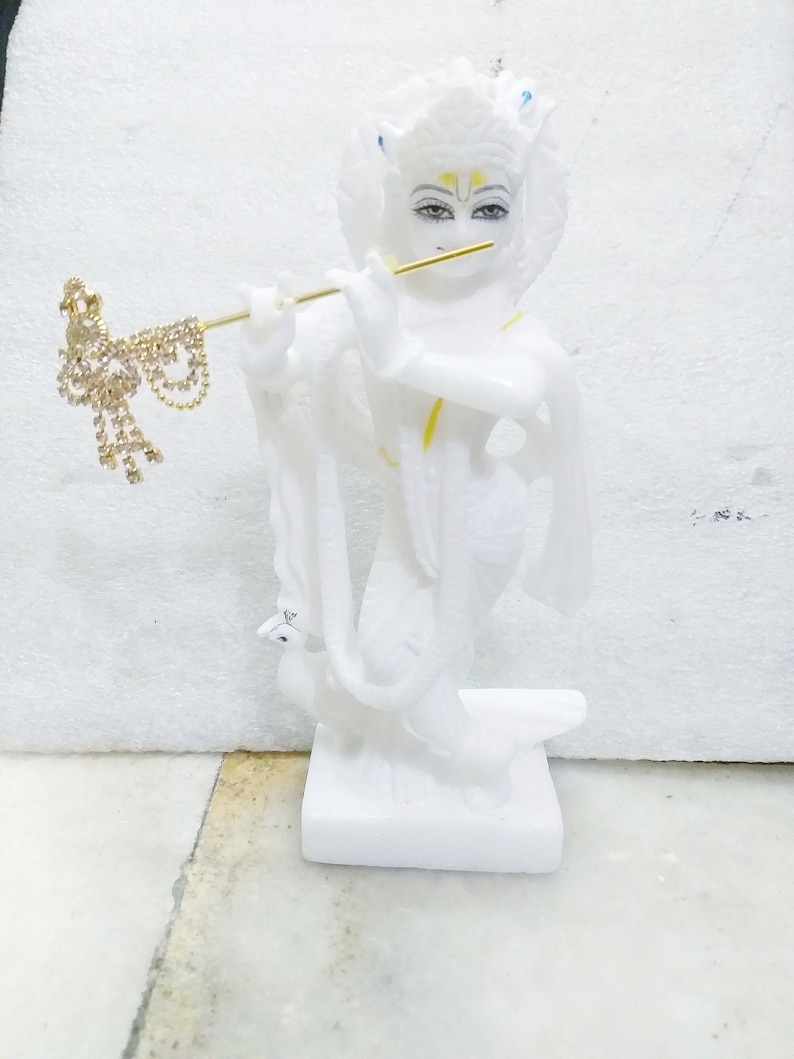 Puede incluir: Estatua de m&aacute;rmol blanco de una deidad hind&uacute; tocando la flauta. La estatua est&aacute; detallada con pintura dorada y tiene una pluma de pavo real en la cabeza.