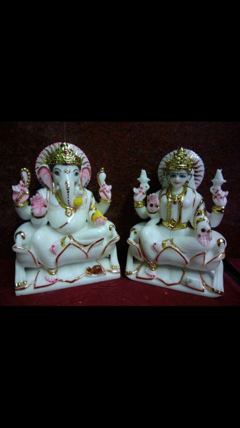May include: White marble statues of Ganesha and Lakshmi, Hindu deities, sitting on lotus thrones. Ganesha is depicted with an elephant head and four arms, while Lakshmi is shown with four arms and a crown. Both statues have gold accents and pink details.