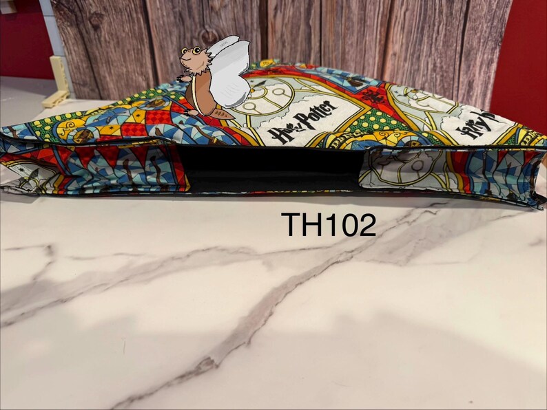 Może przedstawiać: Kolorowa torba typu tote z motywem "Harry Potter" i fantazyjną ilustracją. Torba ma czarne wnętrze, widoczny jest napis "TH102".