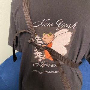 以下が含まれることがあります： 「New York Silkworm」というテキストと、カイコのグラフィックが描かれた黒いTシャツ。黒い調節可能なストラップがシャツの上にかかっています。ウェブサイト「www.nyosilkworms.com」も表示されています。